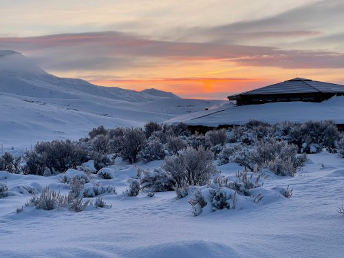 A snowy landscape at sunset, with mountains in the background and a silhouette of a building in the foreground.
