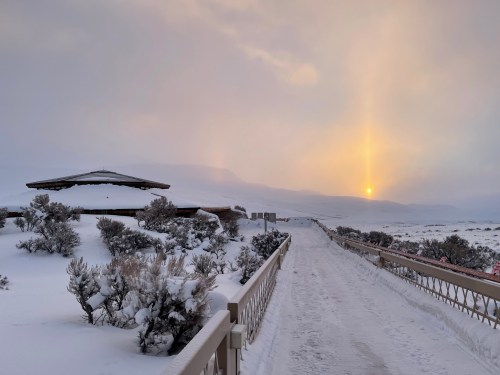 A snowy path leads to a gazebo, with a soft sunset illuminating the mountains in the background.