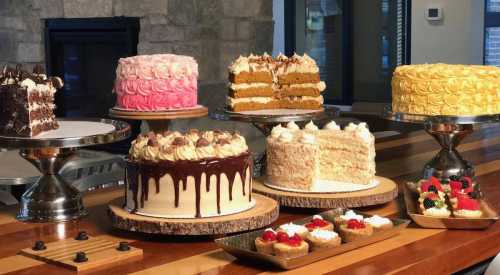 A variety of beautifully decorated cakes and desserts displayed on wooden stands, including chocolate, vanilla, and fruit toppings.