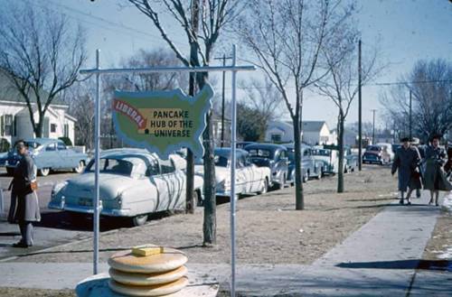 A vintage street scene featuring a sign for "Liberal, Pancake Hub of the Universe" with parked cars and trees.