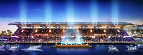 A modern waterfront venue with bright lights, an audience, and boats in the foreground, set against a twilight sky.