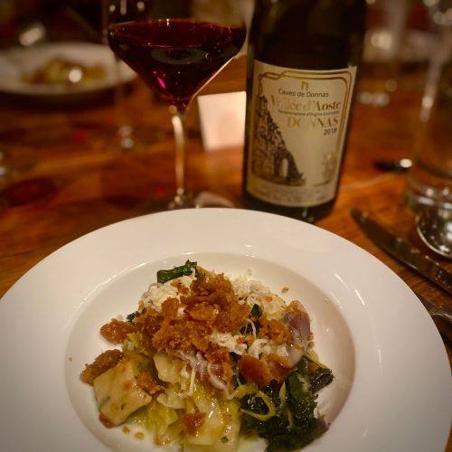 A plate of pasta topped with greens and crispy bits, accompanied by a glass of red wine and a bottle in the background.