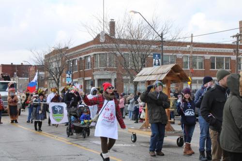 A festive parade scene with participants in costumes, banners, and a decorated cart, set in a city street.