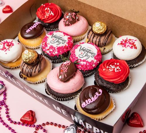 A box of assorted Valentine's Day cupcakes in pink, red, and chocolate, decorated with sprinkles and chocolate treats.