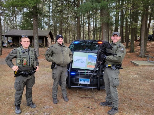 Three law enforcement officers stand by a police vehicle in a wooded area, one holding a dog, with a sign nearby.