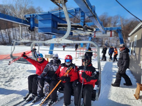 Four skiers on a chairlift, smiling and holding skis, with a snowy landscape and other skiers in the background.