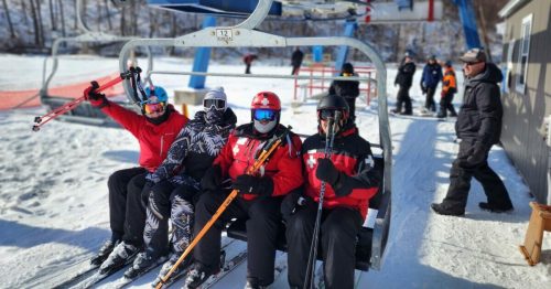 Four skiers on a chairlift, smiling and holding skis, with a snowy landscape and other skiers in the background.