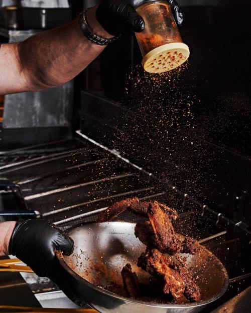 A chef in black gloves sprinkles seasoning over a bowl of crispy chicken wings in a kitchen setting.