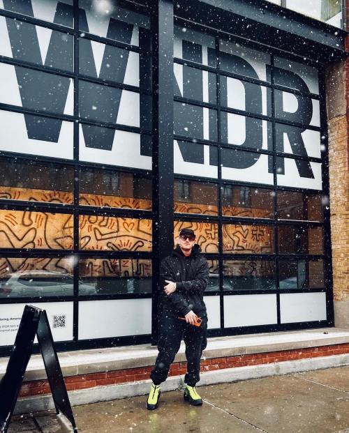 A person stands in front of a large window with "WNDR" displayed, snow falling around them.
