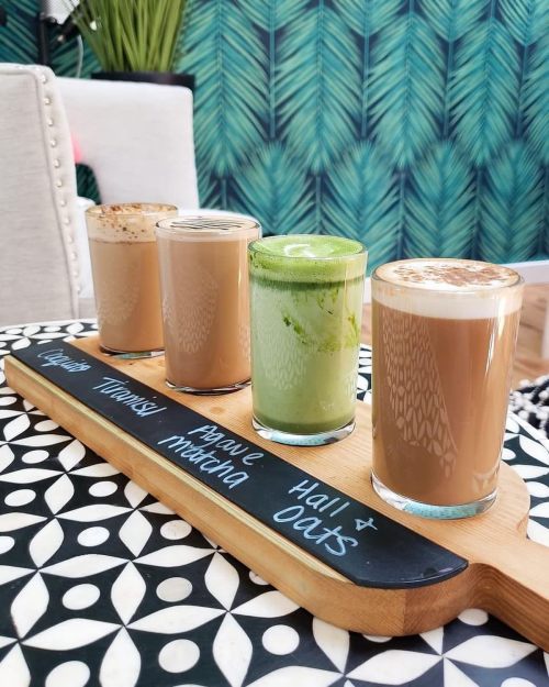 A wooden tray holds four different drinks: two lattes, a green beverage, and a frothy drink, set against a patterned background.