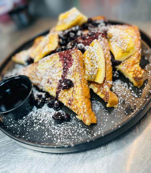 A plate of French toast topped with powdered sugar, banana slices, and dried fruit, served with a small cup of syrup.