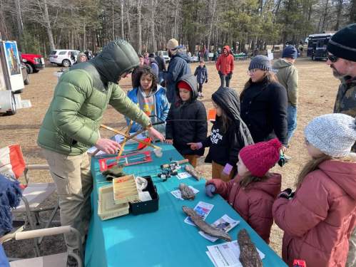 A group of children and adults gather around a table, learning about nature tools from an instructor in a park setting.