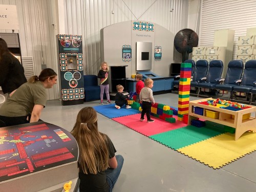 Children play with colorful building blocks in a spacious indoor activity area, with adults supervising nearby.