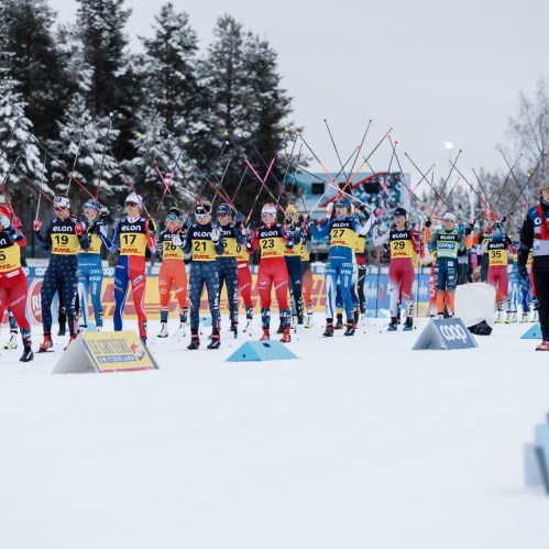 A group of cross-country skiers in colorful uniforms compete in a snowy landscape, raising their poles in unison.
