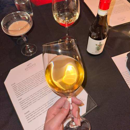 A hand holding a glass of white wine, surrounded by other drinks and menus on a table.