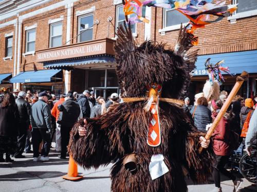 A person in a bear costume stands in front of the Slovenian National Home, surrounded by a crowd during a festive event.