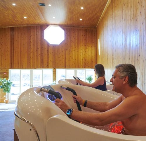 Two people enjoying a spa experience in a wooden room, using hydrotherapy tubs with modern controls.