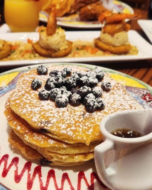 A stack of pancakes topped with blueberries and powdered sugar, served with a small cup of syrup.