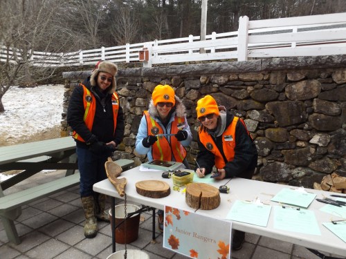 Three people in bright orange vests and yellow hats smile at a table with nature materials, promoting a Junior Rangers program.
