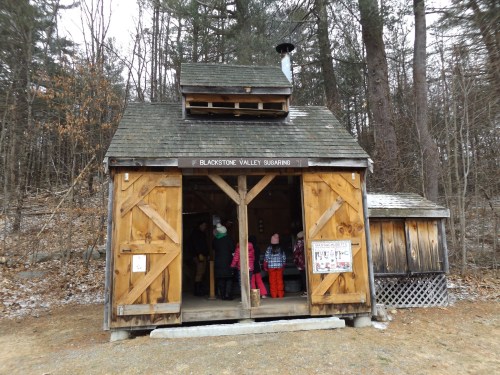 A wooden sugar shack in a forested area, with visitors entering and snow on the ground.
