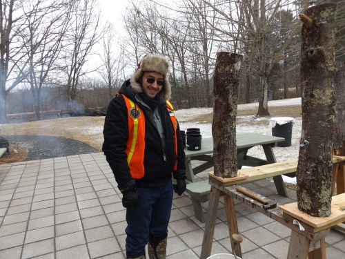 A person in a winter outfit stands next to two logs on a wooden stand, with a snowy landscape in the background.