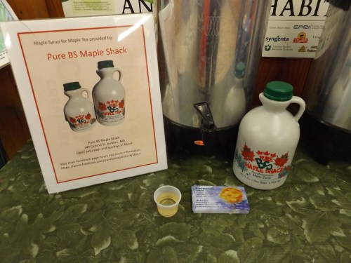 A display featuring a sign for Pure BS Maple Shack, a syrup bottle, a tasting cup, and a business card.