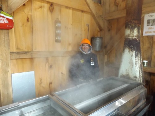 A person in a bright orange hat stands in a wooden cabin, surrounded by steam from a large metal container.