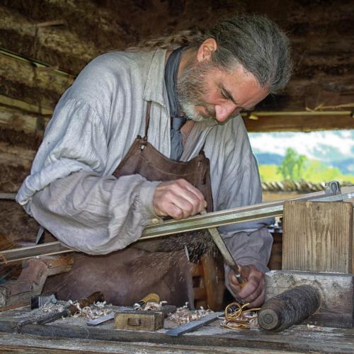 A craftsman works with wood, using a hand tool in a rustic workshop filled with tools and natural light.