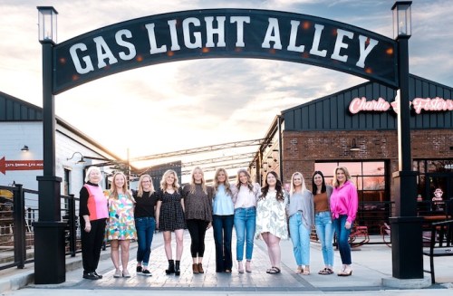 A group of women poses together under the "Gas Light Alley" sign at sunset, with buildings in the background.