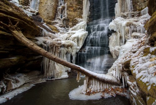 A frozen waterfall surrounded by icy rocks and a fallen tree, creating a serene winter landscape.