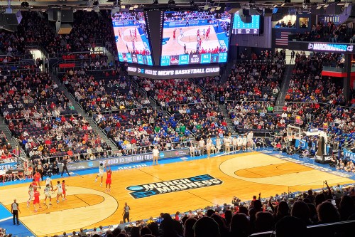A packed arena during a college basketball game, featuring large screens and teams on the court.