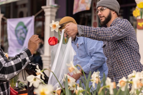 A man in a beanie hands a bag of flowers to another person at a market, surrounded by blooming plants.