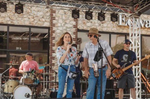 A woman and a man speak on stage at a music event, with a band and audience in the background.