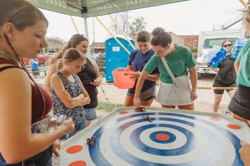 A group of young women watches as one girl plays a game on a circular target board under a tent.