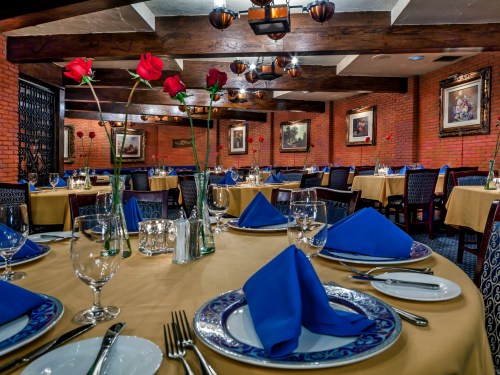 Elegant restaurant interior with tables set for dining, featuring blue napkins and red roses in vases. Warm, inviting atmosphere.
