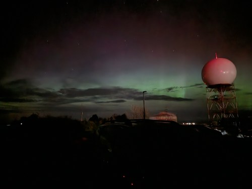 A night sky illuminated by auroras, with a radar dome and silhouettes of trees in the foreground.