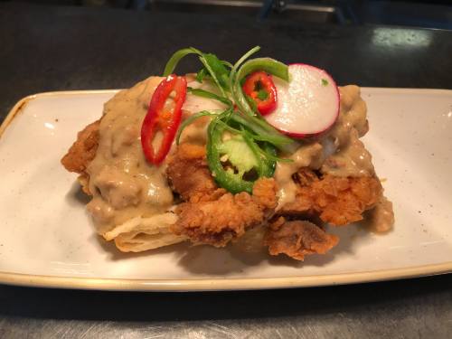 A plate of fried chicken on a biscuit, topped with creamy gravy, sliced jalapeños, radishes, and red chili peppers.