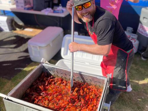A smiling man in an apron stirs a large pot of crawfish at an outdoor event.