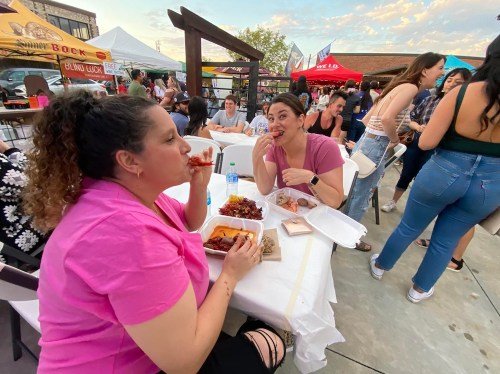 Two women enjoy food at an outdoor event, surrounded by people and colorful tents in a lively atmosphere.