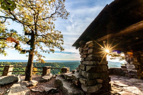 A scenic view from a stone structure, with a tree and sunlight shining over a lush valley and distant hills.