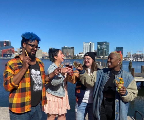 Four friends celebrate by the water, raising drinks in a toast against a sunny city skyline backdrop.