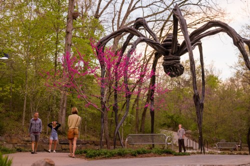 A large spider sculpture towers over visitors near a blooming pink tree in a wooded area.