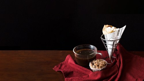 A cup of coffee, a muffin, and a cone with a pastry, arranged on a wooden table with a red cloth.