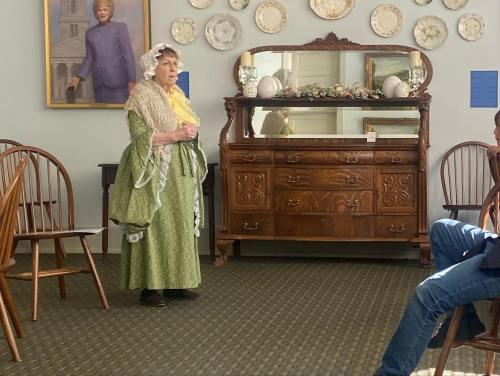 An elderly woman in period costume stands in a room with wooden furniture and decorative plates on the wall.
