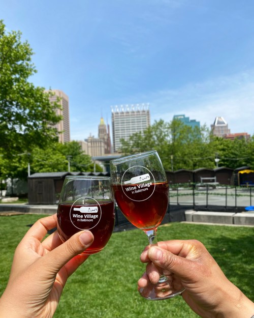 Two hands hold glasses of red wine, toasting against a city skyline and clear blue sky in a park setting.