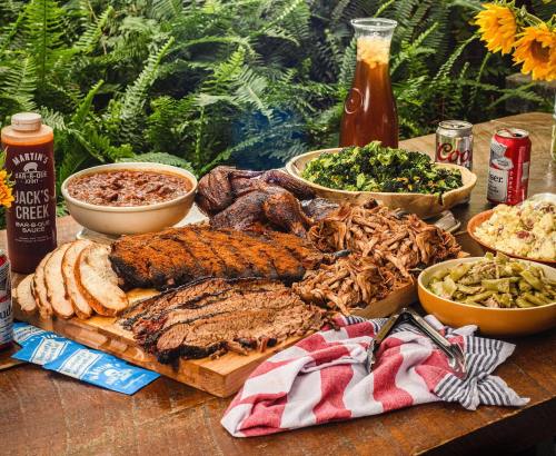A spread of barbecue featuring brisket, pulled pork, sides, sauces, and drinks, set on a rustic table with greenery.