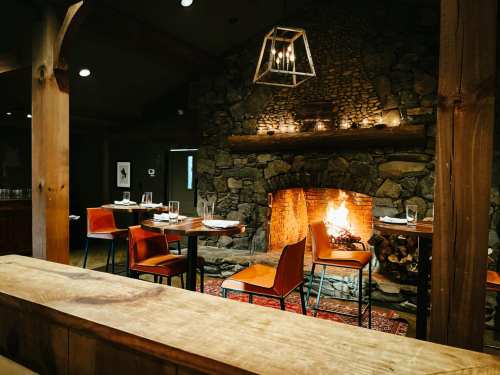 Cozy restaurant interior with a stone fireplace, wooden beams, and tables set for dining. Warm, inviting atmosphere.