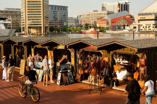 A waterfront market with wooden stalls selling bags, people browsing, and a cyclist passing by in the foreground.