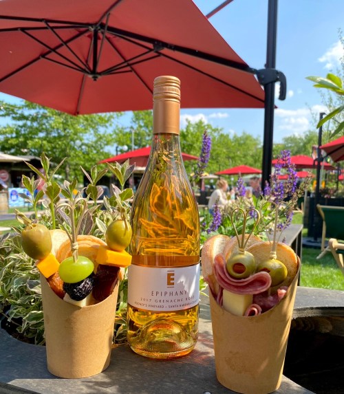 A bottle of Epiphany rosé wine surrounded by two cups of assorted fruits, cheeses, and meats under red umbrellas.