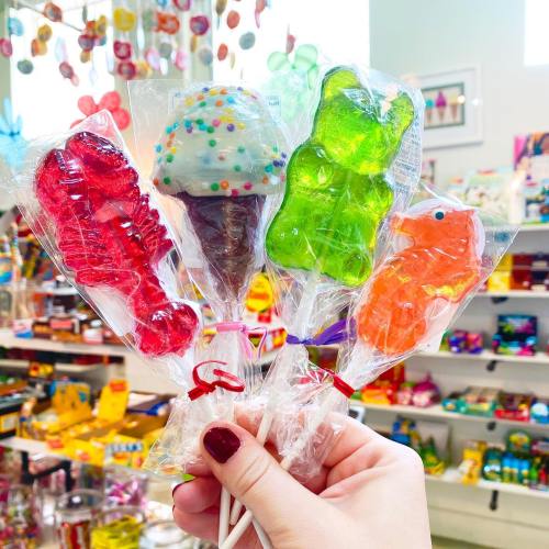 A hand holding colorful lollipops shaped like animals and ice cream, wrapped in clear plastic, against a bright candy store backdrop.